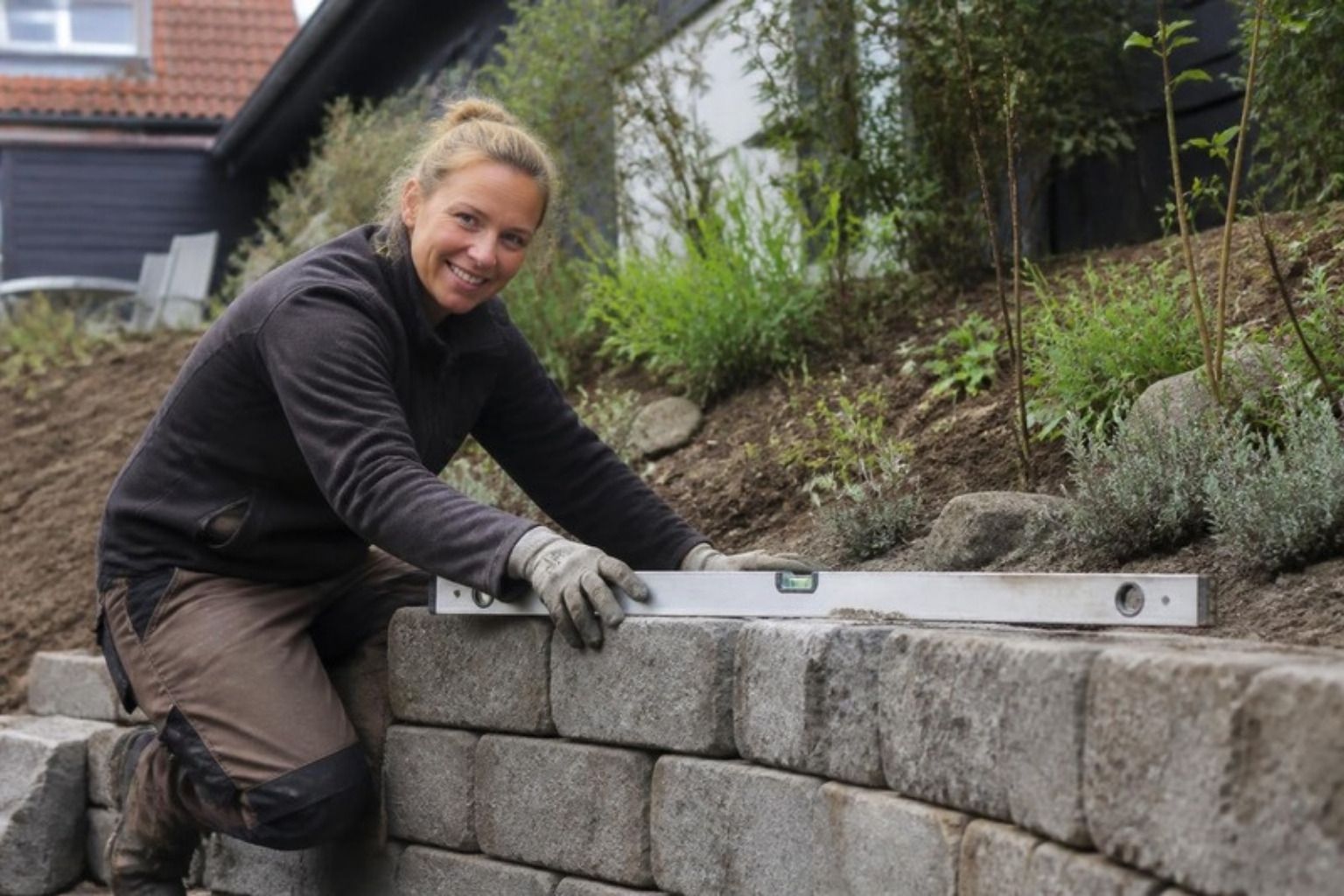Smilende kvinde bygger en lav støttemur af grå betonblokke og tjekker den med vaterpas