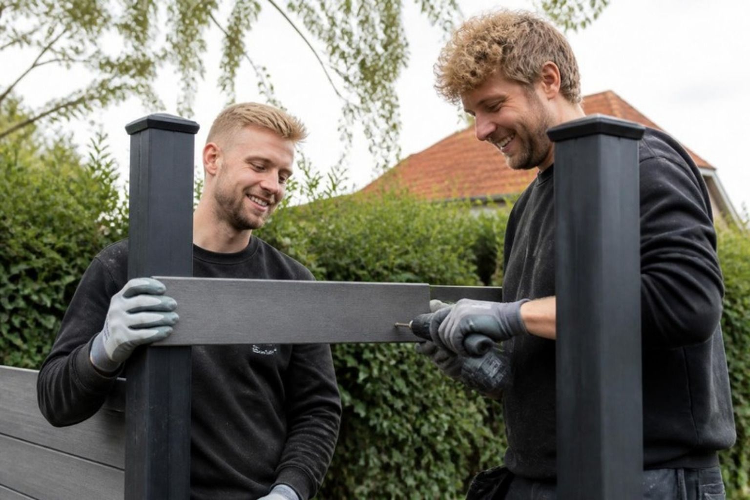 To smilende mænd monterer et gråt komposithegnsbræt mellem mørke stolper